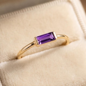 Peut inclure: Une bague en or ornée d'une unique pierre précieuse améthyste violette de taille baguette rectangulaire, sertie horizontalement dans une monture en métal lisse. L'anneau est fin et poli, présentant un design épuré mettant en valeur la pierre.