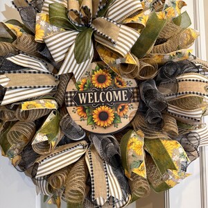 May include: A decorative wreath featuring a central circular sign with the text WELCOME surrounded by sunflowers. The wreath is constructed from layers of mesh and fabric ribbons in shades of olive green, tan, black, and white with sunflower patterns.