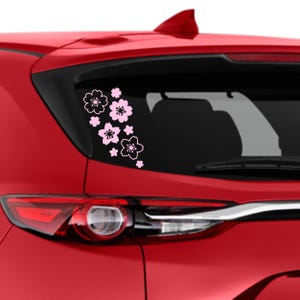 May include: A vinyl decal featuring a cluster of light pink cherry blossom flowers with white outlines applied to the rear window of a red vehicle. The design consists of several blossoms of varying sizes arranged in a cascading floral pattern.