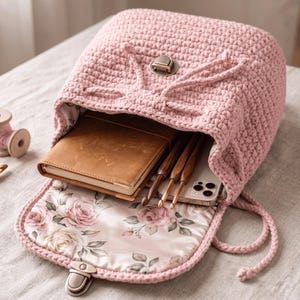 May include: A light pink crochet backpack with a drawstring closure and a metal clasp. The interior is lined with a floral rose pattern fabric and contains a brown notebook, three wooden crochet hooks, and a smartphone. Text reads Crochet Rose Backpack Pattern and Instant PDF Download.