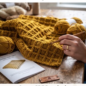 Pode incluir: Uma manta de crochê amarelo-mostarda com textura de ponto waffle, acompanhada de novelos de lã correspondentes, uma agulha de crochê de madeira, um documento de padrão impresso intitulado Premium Pattern e uma etiqueta de couro Handmade Pure Wool.