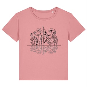 Könnte beinhalten: Ein altrosa T-Shirt mit kurzen Ärmeln und einer schwarzen Strichzeichnung verschiedener Wildblumen und Gräser über einer horizontalen Linie, wobei sich unterhalb der Linie eine gespiegelte Reflexion des Blumenmotivs befindet.
