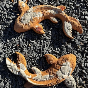 May include: Two koi fish sculptures crafted from terracotta-colored material, featuring intricate, raised swirl patterns across their bodies. The fish display a dual-tone aesthetic with natural orange-brown hues and contrasting white textured accents.