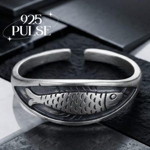 May include: An open-ended silver ring featuring a detailed fish motif embossed on a dark, recessed background. The metal has a matte, oxidised finish. The text 925 PULSE is visible in the upper left corner of the image, indicating the material quality.