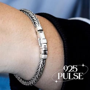 Könnte beinhalten: Ein silbernes Armband aus geflochtener Kette mit einem polierten, rechteckigen Verschlussmechanismus. Die Metalloberfläche weist ein strukturiertes, geflochtenes Muster auf. Der Text 925 PULSE ist in der unteren rechten Ecke zu sehen.