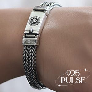 May include: A silver bracelet featuring a woven chain design and a rectangular clasp with a circular emblem containing a symbol. The clasp is decorated with a geometric Greek key pattern. The text 925 PULSE is visible in the bottom right corner.