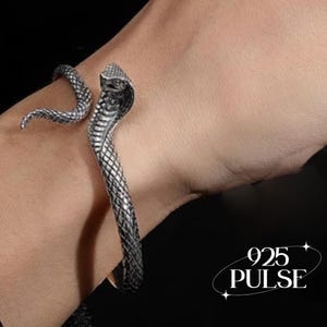 May include: A silver-toned metal cuff bracelet shaped like a cobra snake with detailed scale textures. The snake head is positioned at one end of the open band, while the tail curves around the other side. The text 925 PULSE is visible.