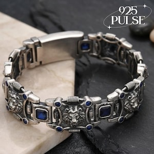 Puede incluir: Una pulsera de eslabones de metal plateado con motivos repetitivos de cabezas de león y piedras azules de corte cuadrado. El diseño incluye pequeños acentos azules circulares y detalles de metal estriado. El texto 925 PULSE es visible arriba a la derecha.
