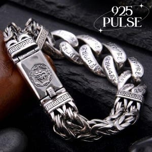 Puede incluir: Una pulsera de cadena de plata con eslabones gruesos entrelazados grabados con caracteres de escritura repetidos. El cierre rectangular muestra un emblema circular con un símbolo intrincado y patrones geométricos. El texto 925 PULSE es visible.