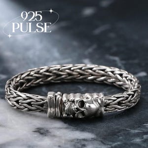 May include: A silver bracelet featuring a woven chain design and a central clasp shaped like a human skull. The text 925 PULSE is visible in the upper left corner. The metal has a polished finish with dark oxidized accents in the recessed areas.