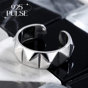 May include: A silver-coloured open band ring featuring a series of raised triangular spikes around the exterior surface. The interior of the band is stamped with the text 925. The text 925 PULSE is visible in the upper left corner of the image.