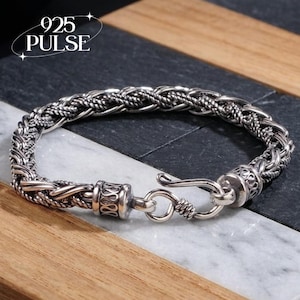 Peut inclure: Un bracelet en argent présentant un design de chaîne tressée épaisse semblable à une corde avec des détails oxydés. Le bracelet comprend un mécanisme de fermoir à crochet et un embout cylindrique avec des motifs gravés. Le texte 925 PULSE est visible.