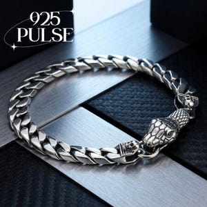 May include: A silver-toned curb chain bracelet featuring a detailed snake head clasp and small skull accents. The text 925 PULSE is visible in the upper left corner. The bracelet is constructed from polished metal links with a textured snake scale pattern.