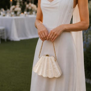 Peut inclure: Une robe blanche sans manches avec un décolleté drapé et un long pan de tissu transparent sur l'épaule. La robe est accompagnée d'une pochette rigide couleur crème en forme de coquillage avec une chaîne dorée et un petit fermoir doré.