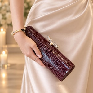 May include: A long, cylindrical clutch bag in a deep burgundy color with a textured crocodile skin pattern. The bag features a metallic silver-toned clasp on the top edge and is held against a smooth, champagne-colored satin fabric garment.