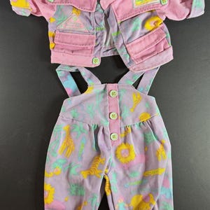 May include: A pink jacket and matching lavender overalls set featuring a colorful animal print of giraffes, lions, and elephants. The jacket has light green buttons and a Cabbage Patch Kids patch. The overalls have three light green buttons.
