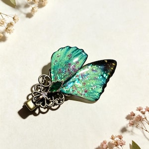 May include: A hair clip featuring a butterfly-shaped ornament with iridescent green and teal wings containing embedded glitter and purple flecks, attached to an ornate bronze-toned metal base with a small green center stone.