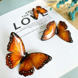 May include: Three glossy butterfly ornaments featuring vibrant orange and brown wings with black edges and white spots. The butterflies are arranged on a white surface printed with black text that reads AROUND THE CORNER LOVE and other phrases.
