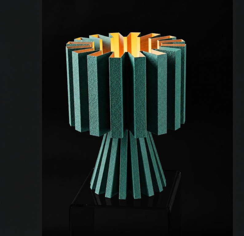 Puede incluir: Una l&aacute;mpara de mesa color verde azulado con una pantalla cil&iacute;ndrica plisada y texturizada y una base plisada a juego. El interior de la pantalla presenta un forro de color claro y c&aacute;lido que contrasta con el exterior verde azulado oscuro.