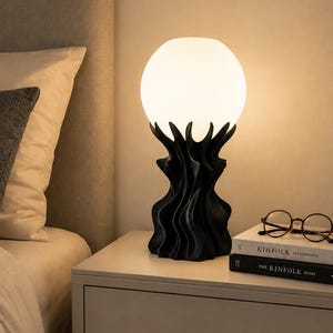 Puede incluir: Una lámpara de mesa con una pantalla esférica de cristal blanco sobre una base escultórica negra con crestas verticales fluidas, orgánicas y onduladas. La lámpara emite un brillo cálido y suave, iluminando el área circundante con luz tenue.