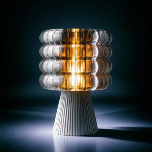 Puede incluir: Una lámpara de mesa con una pantalla cilíndrica de vidrio acanalado que emite una luz cálida de tono ámbar. La base es una estructura cónica blanca y sólida con estrías verticales que sostiene la parte superior de vidrio translúcido.