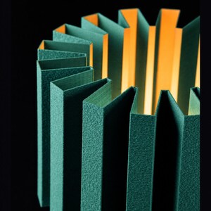Puede incluir: Una pantalla de l&aacute;mpara cil&iacute;ndrica con un exterior plisado en un color verde bosque profundo con un acabado texturizado. El interior de la pantalla es de un naranja brillante y c&aacute;lido, creando un fuerte contraste al iluminar los pliegues.