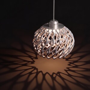 May include: A spherical pendant light fixture featuring a complex, interconnected lattice structure in a metallic silver finish. The lamp is suspended by a thin cord and emits a warm glow, casting intricate, repeating shadow patterns below it.