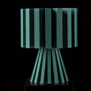 Puede incluir: Una l&aacute;mpara de mesa de color verde azulado con una pantalla cil&iacute;ndrica compuesta por paneles rectangulares verticales espaciados uniformemente. La base es del mismo color, c&oacute;nica y formada por paneles verticales sobre un soporte cuadrado negro.