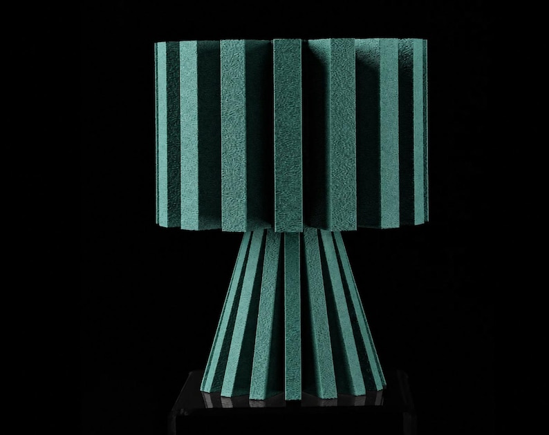 Puede incluir: Una l&aacute;mpara de mesa de color verde azulado con una pantalla cil&iacute;ndrica compuesta por paneles rectangulares verticales espaciados uniformemente. La base es del mismo color, c&oacute;nica y formada por paneles verticales sobre un soporte cuadrado negro.