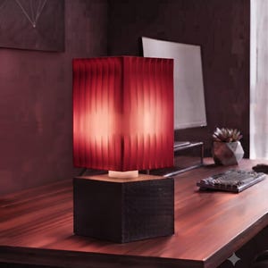 Puede incluir: Una lámpara de mesa con una base cuadrada de madera oscura y una pantalla rectangular con nervaduras verticales en un tono rojo intenso. La lámpara emite un brillo suave y cálido desde su interior, resaltando la textura de la pantalla.