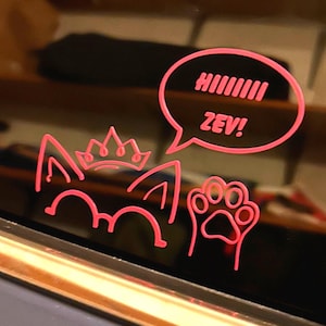 May include: A bright pink vinyl decal applied to a dark surface, featuring a stylized cat head with pointed ears, a small crown, a paw print, and a speech bubble containing the text HIIIIIII ZEV! in a bold, sans-serif font.