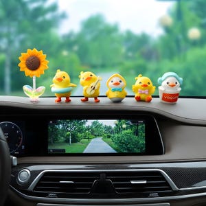 May include: A set of six decorative dashboard ornaments featuring a yellow sunflower on a spring, four yellow ducklings in various poses including one with a guitar, and a small white figure wearing blue headphones inside a red yogurt cup.