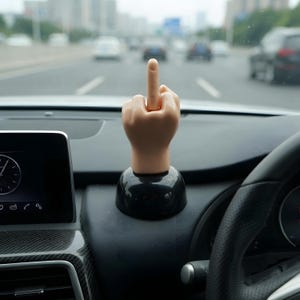 May include: A light-skinned plastic hand figurine displaying a middle finger gesture, mounted on a smooth, rounded black base. The object is positioned on the dashboard of a vehicle, with the fingers curled and the middle finger extended upward.