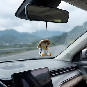 Op de afbeelding: Een wit pinguïnfiguurtje hangend aan een zwart koord bevestigd aan een achteruitkijkspiegel. De pinguïn draagt een strooien hoed, een bril met zwart montuur en een gele sjaal, met oranje voeten naar voren gestrekt in een zittende houding.