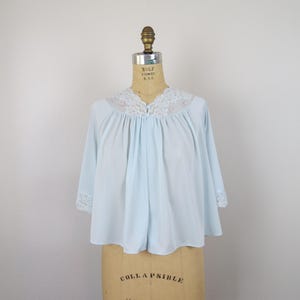 May include: A light blue bed jacket featuring a wide white lace collar with small pink floral accents. The garment has three-quarter length sleeves finished with matching white lace cuffs and a single button closure at the center neckline.