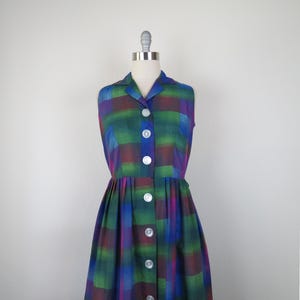 May include: A sleeveless dress featuring a plaid pattern in shades of green, blue, and purple. The garment has a collared neckline, a button-down front with six visible white buttons, and a pleated skirt that falls from the waistline.