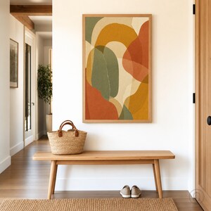 Può includere: Una stampa artistica da parete rettangolare con forme organiche astratte in tonalità verde oliva, giallo senape e arancione terracotta su sfondo crema, incorniciata in legno chiaro sopra una panca in legno con un cesto intrecciato e scarpe.