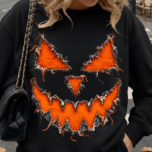 Könnte beinhalten: Ein schwarzes Langarm-Sweatshirt mit einer großen, zentralen Grafik eines Kürbisgesichts. Das Design besteht aus orangefarbenen Stoffaufnähern mit ausgefransten, abgenutzten Kanten, die auf den schwarzen Stoff genäht wurden, um zwei dreieckige Augen, eine kleine dreieckige Nase und ein breites, gezacktes Grinsen zu bilden.
