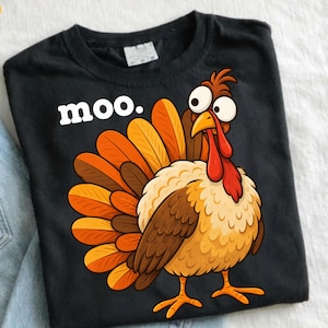 Op de afbeelding: Een zwart t-shirt met een cartoonkalkoen met grote, verbaasde ogen en een waaierstaart van oranje en bruine veren. Het woord "moo." is in witte, vette kleine letters gedrukt aan de linkerkant van de kop van de kalkoen op de borst.