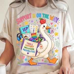 May include: A beige t-shirt featuring a colorful graphic of a white goose carrying a tote bag filled with school and therapy supplies. The text reads SUPPORT IS ON THE WAY, TOOLS SUPPORT INCLUSION POSSIBILITIES, KIND PEOPLE BRIGHTER FUTURES, and DIFFERENT MINDS BRIGHTER WORLDS.