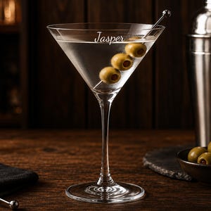 May include: A clear glass martini glass featuring the name Jasper etched on the rim. The glass contains a clear liquid and a metal cocktail pick skewering three green olives. A stainless steel cocktail shaker sits to the right of the glass.