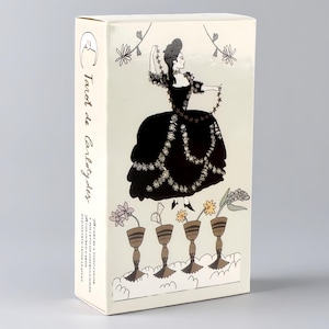 May include: A rectangular tarot card deck box in cream with black line art. The front features a figure in a black gown holding a star garland above four chalices containing flowers. The side text reads Tarot de Carlotydes and 78 coloured cards.