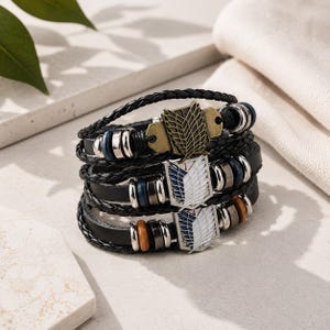 May include: A stack of three black braided leather bracelets featuring metallic charms with wing designs. The charms include a bronze-toned emblem and two silver-toned emblems with blue and white details, accented by silver, blue, and brown beads.