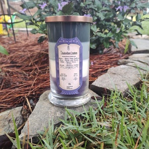 May include: A three-wick candle in a clear glass jar with a copper lid. The candle is layered with black, gray, and purple wax. The label reads "Shadowflame Creations" and lists the scents: Black - Black Violet Bergamot, Gray - Suede and Fir Needle, White - Spiced Honey, Purple - Blueberry Cobbler. The candle has a burn time of 120-130 hours and is 17.5 oz.