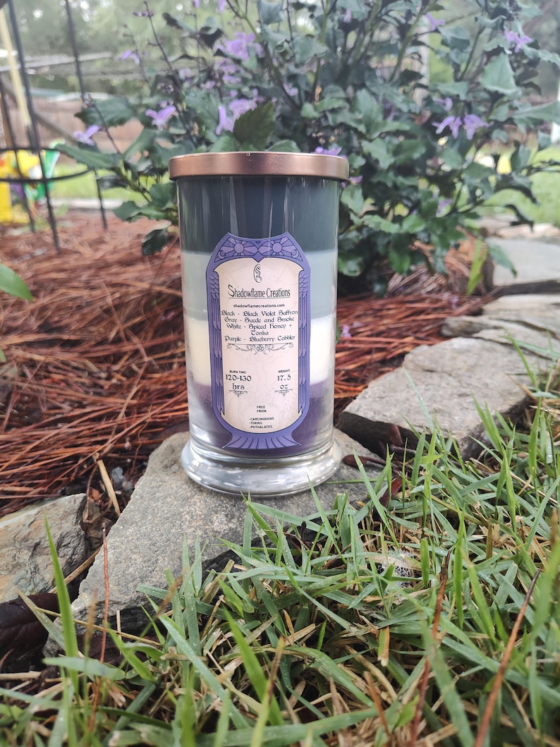 May include: A three-wick candle in a clear glass jar with a copper lid. The candle is layered with black, gray, and purple wax. The label reads "Shadowflame Creations" and lists the scents: Black - Black Violet Bergamot, Gray - Suede and Fir Needle, White - Spiced Honey, Purple - Blueberry Cobbler. The candle has a burn time of 120-130 hours and is 17.5 oz.