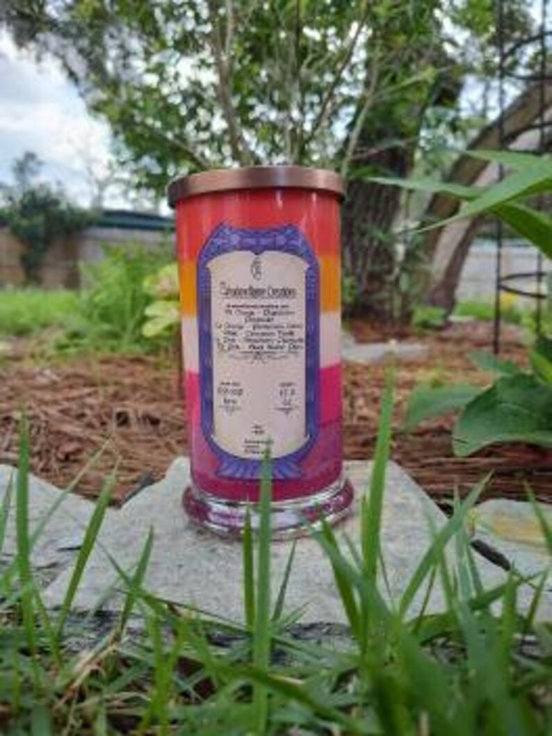Op de afbeelding: Een meerkleurige kaars in een glazen pot met een koperen deksel. De kaars is gelaagd met rode, oranje, gele en paarse was. Het etiket op de pot vermeldt "Southern Charm Creations" en geeft de geur van de kaars aan als "Oranje, Kaneel en Vanille".