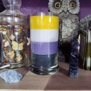 May include: A tall, cylindrical candle with four layers of wax in the colors of the non-binary pride flag: black, purple, white, and yellow. The candle is in a clear glass jar with a gold rim.