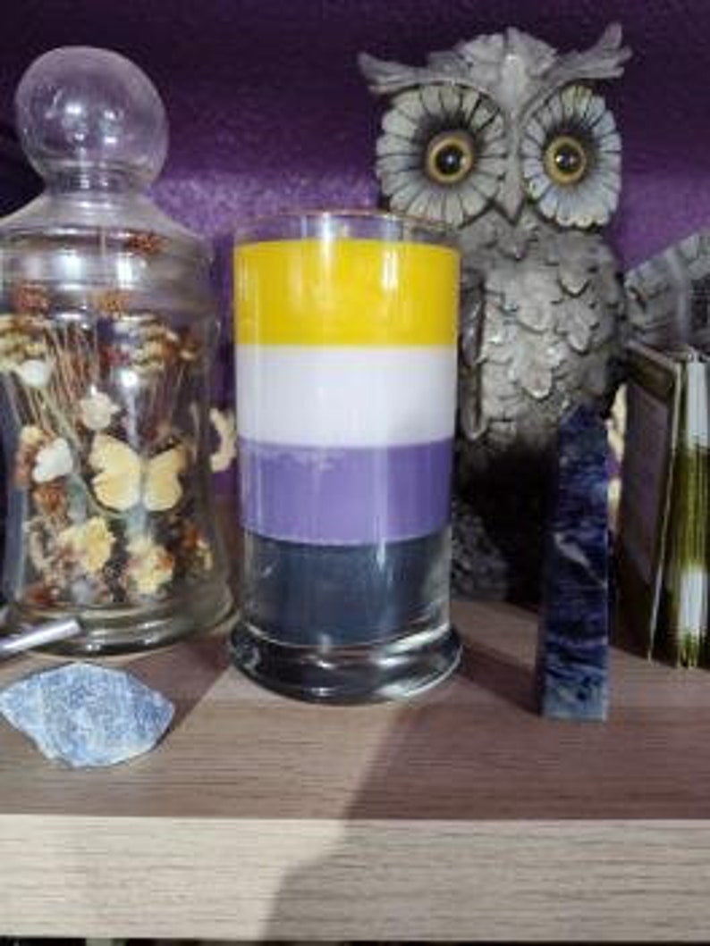 May include: A tall, cylindrical candle with four layers of wax in the colors of the non-binary pride flag: black, purple, white, and yellow. The candle is in a clear glass jar with a gold rim.