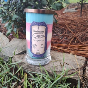 May include: A layered candle in a glass jar with a blue, pink, and white color scheme. The candle is labeled "Shadowflame Creations" and lists the scents "Acqua Di Gio", "Peachos + Cream", "Green Tea", and "Lemongrass". The candle has a burn time of 120-130 hours and weighs 17.5 ounces.