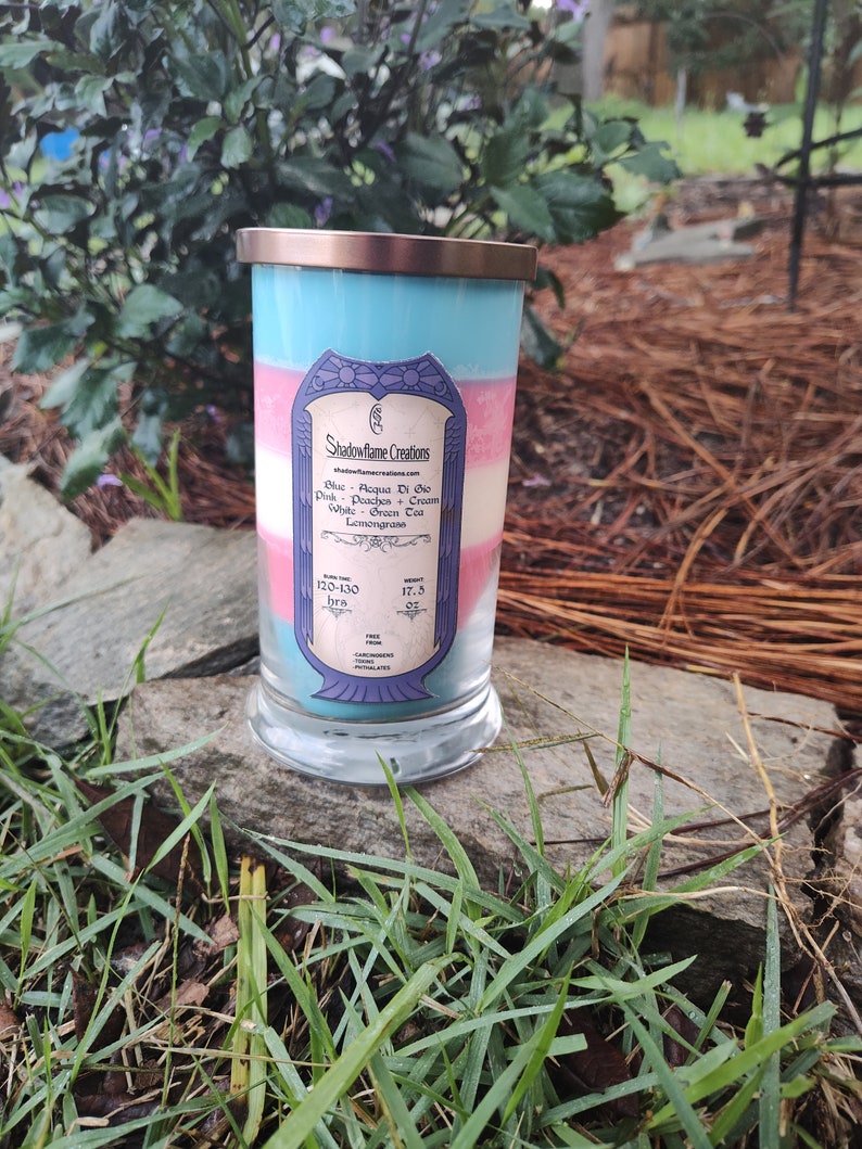May include: A layered candle in a glass jar with a blue, pink, and white color scheme. The candle is labeled "Shadowflame Creations" and lists the scents "Acqua Di Gio", "Peachos + Cream", "Green Tea", and "Lemongrass". The candle has a burn time of 120-130 hours and weighs 17.5 ounces.